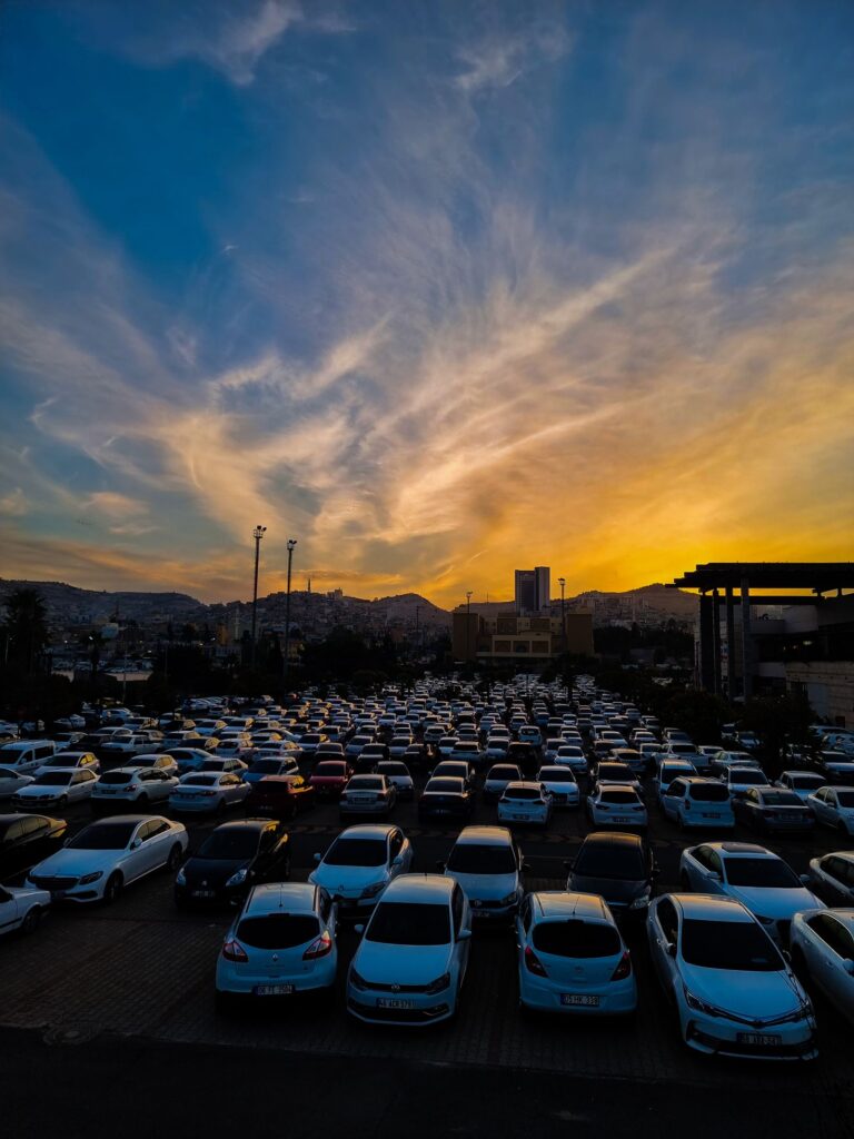 Rows of parked cars on an outdoor lot at sunset with city silhouettes behind